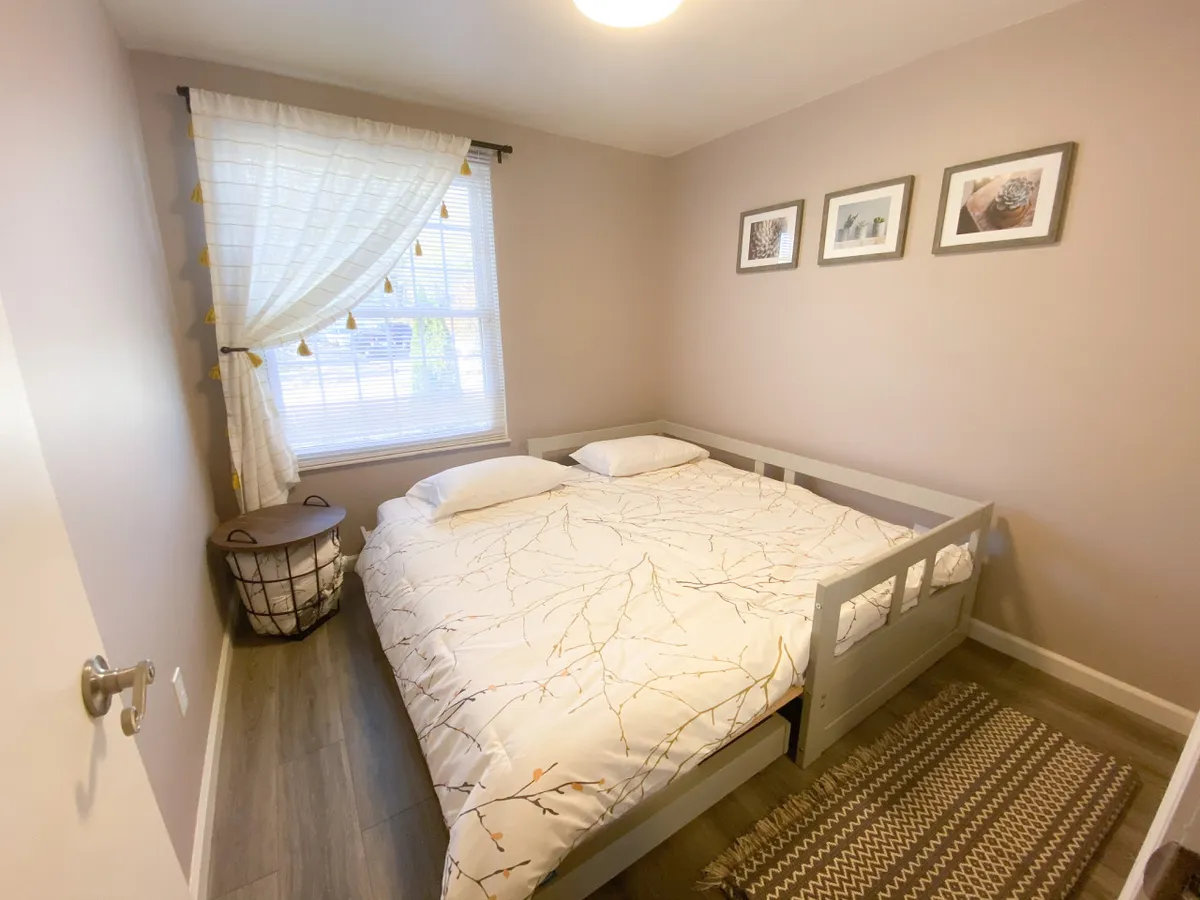 A tranquil bedroom scene with a comfortable bed, sheer-curtained window allowing natural light, framed artwork, and a woven basket, suggesting a peaceful space for rest and family downtime.