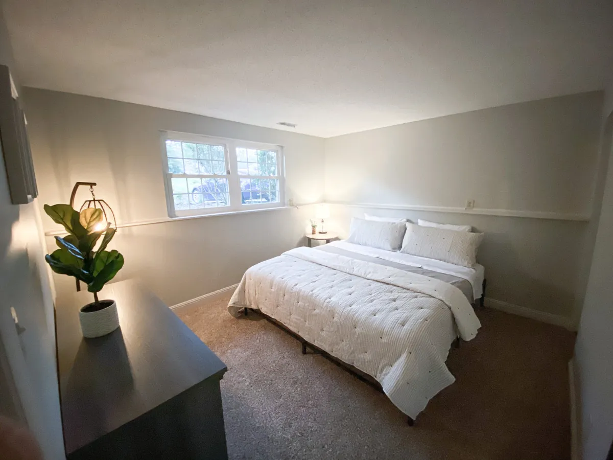 A serene bedroom setup with a bed dressed in white linens, a nightstand with a lamp, and a potted plant. The room is bathed in natural light, evoking a sense of calm and restfulness—perfect for unwinding after a long day.