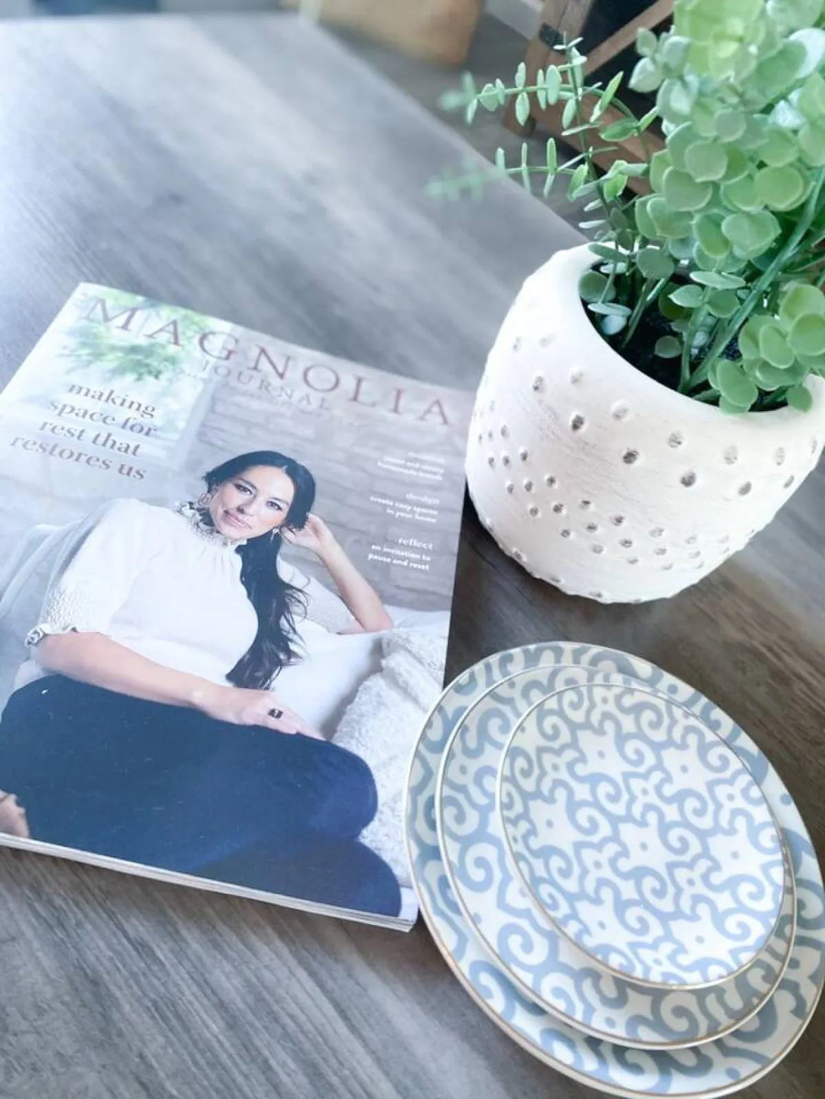 A tastefully decorated table with Magnolia Journal magazine, decorative plates, and a potted plant. The setup alludes to creative brainstorming, relaxing breaks, or entertaining guests, infusing the space with a welcoming, home-like atmosphere.