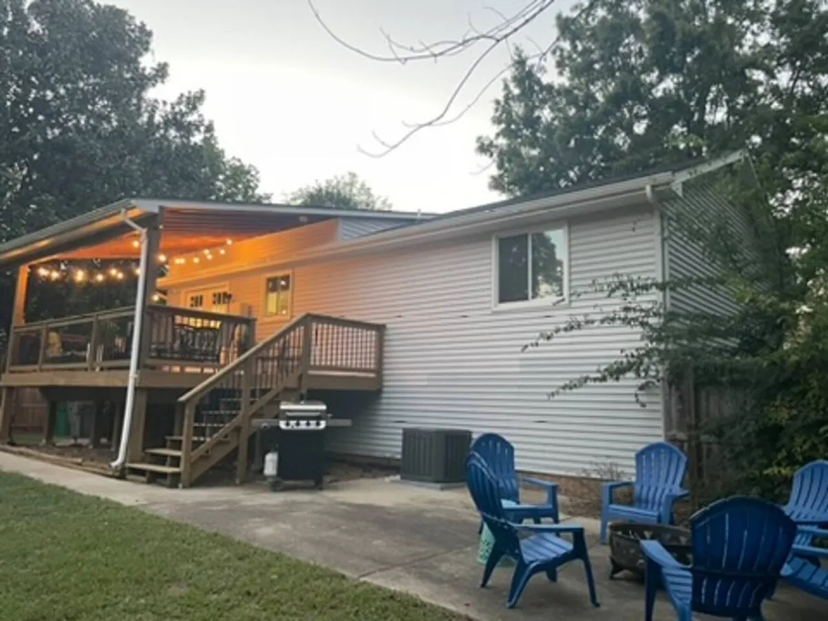 This exterior image showcases a spacious deck adorned with string lights and blue Adirondack chairs, creating an inviting atmosphere. A grill is prominently positioned, suggesting opportunities for outdoor cooking. The overall scene captures a lively yet relaxed area designed for social gatherings and outdoor dining.