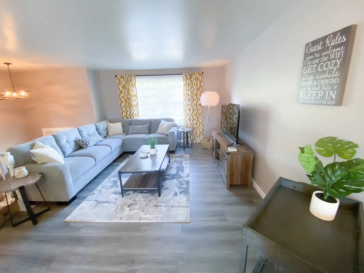 A cozy living room with a comfortable sofa, coffee table, curtains framing a window, decorative plants, and a TV. The space is ideal for socializing, relaxing after a day out, or enjoying shared activities like watching TV or having drinks.