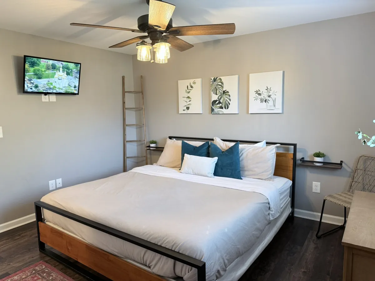 The hero image showcases a beautifully styled bedroom with a plush bed adorned with decorative pillows, elegant wall art, a sleek television, and a modern ladder shelf. The space feels bright, inviting, and designed for comfort—perfect for unwinding after a day exploring Charlotte.