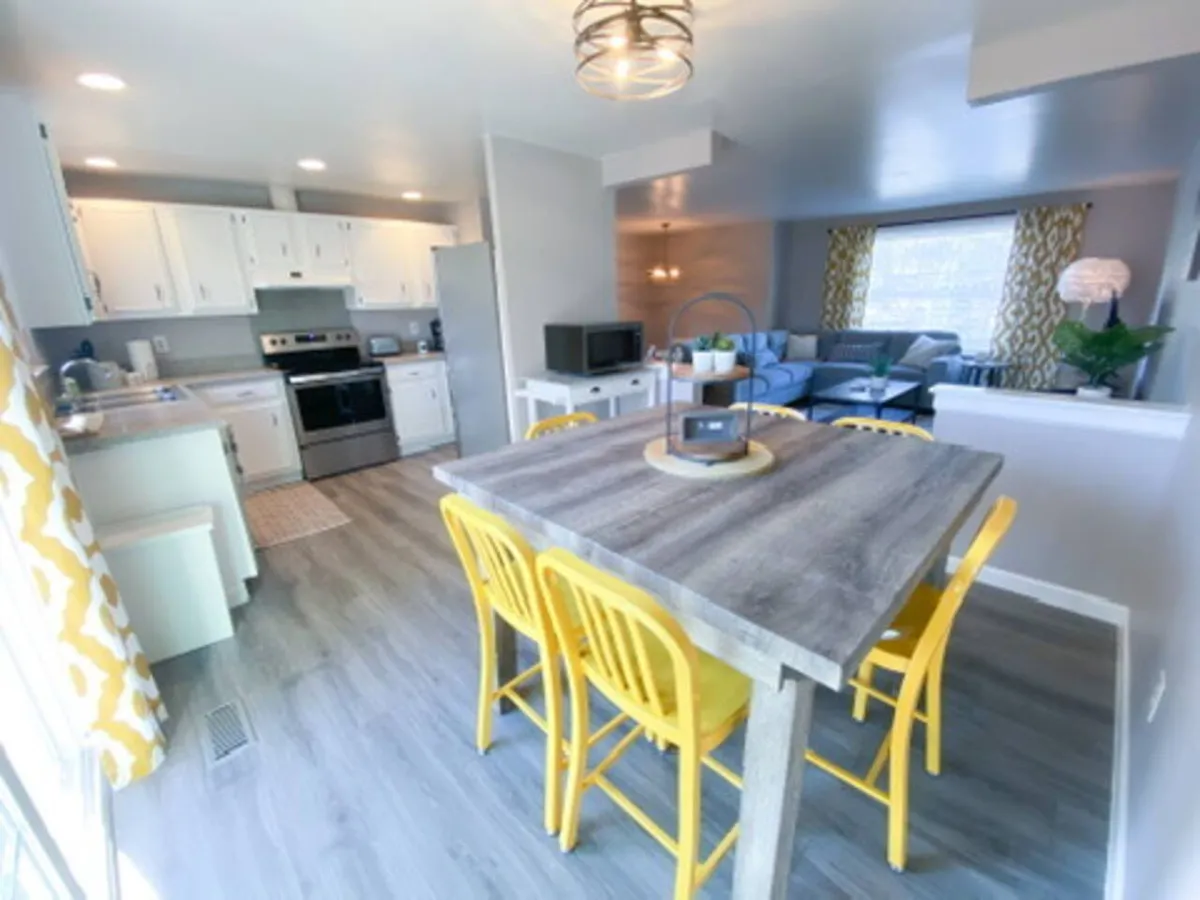 A modern kitchen and dining area featuring yellow chairs at a wooden table, sleek kitchen appliances, and an open view into the cozy living room. The space feels cheerful and inviting, designed for both cooking and socializing.