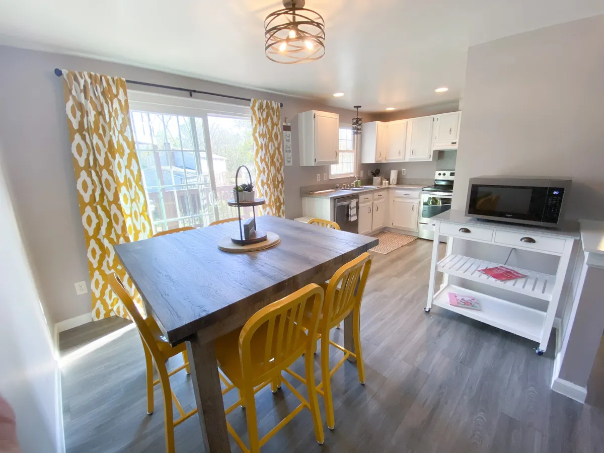 This image showcases a large wooden dining table surrounded by bright yellow chairs, colorful curtains framing the space, and modern kitchen appliances visible in the background. The scene exudes a welcoming, communal atmosphere ideal for shared meals and gatherings.