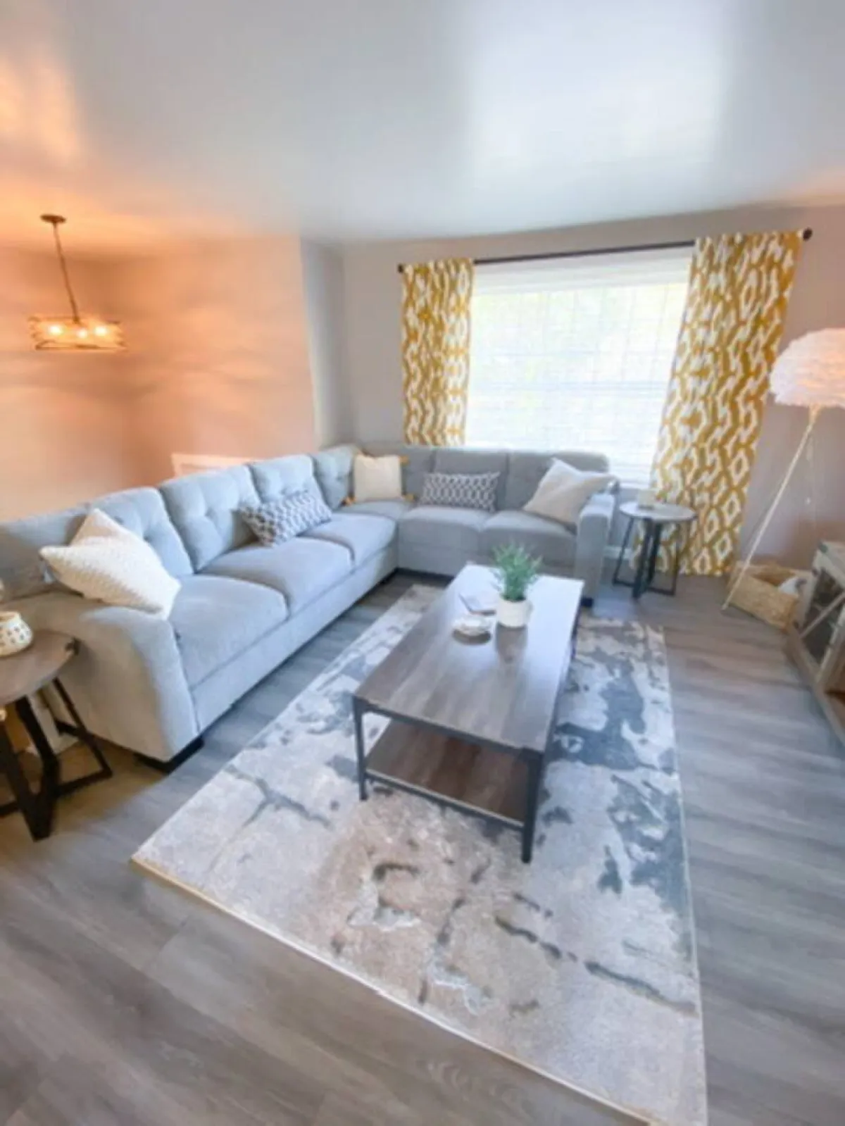 A cozy, inviting living room featuring a gray sectional sofa adorned with decorative pillows, a wooden coffee table at the center, and cheerful yellow patterned curtains. The space exudes warmth and is perfect for relaxing, reading, or hosting intimate gatherings.