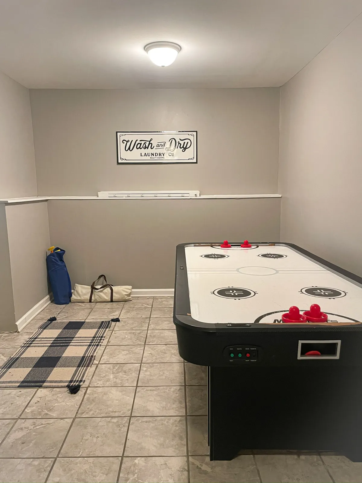 A playful, light-hearted area with an air hockey table positioned on a decorative rug. A 'Wash and Dry' sign hints at the multifunctionality of the space, but the primary focus is on the game table, ideal for friendly competition and shared laughter.
