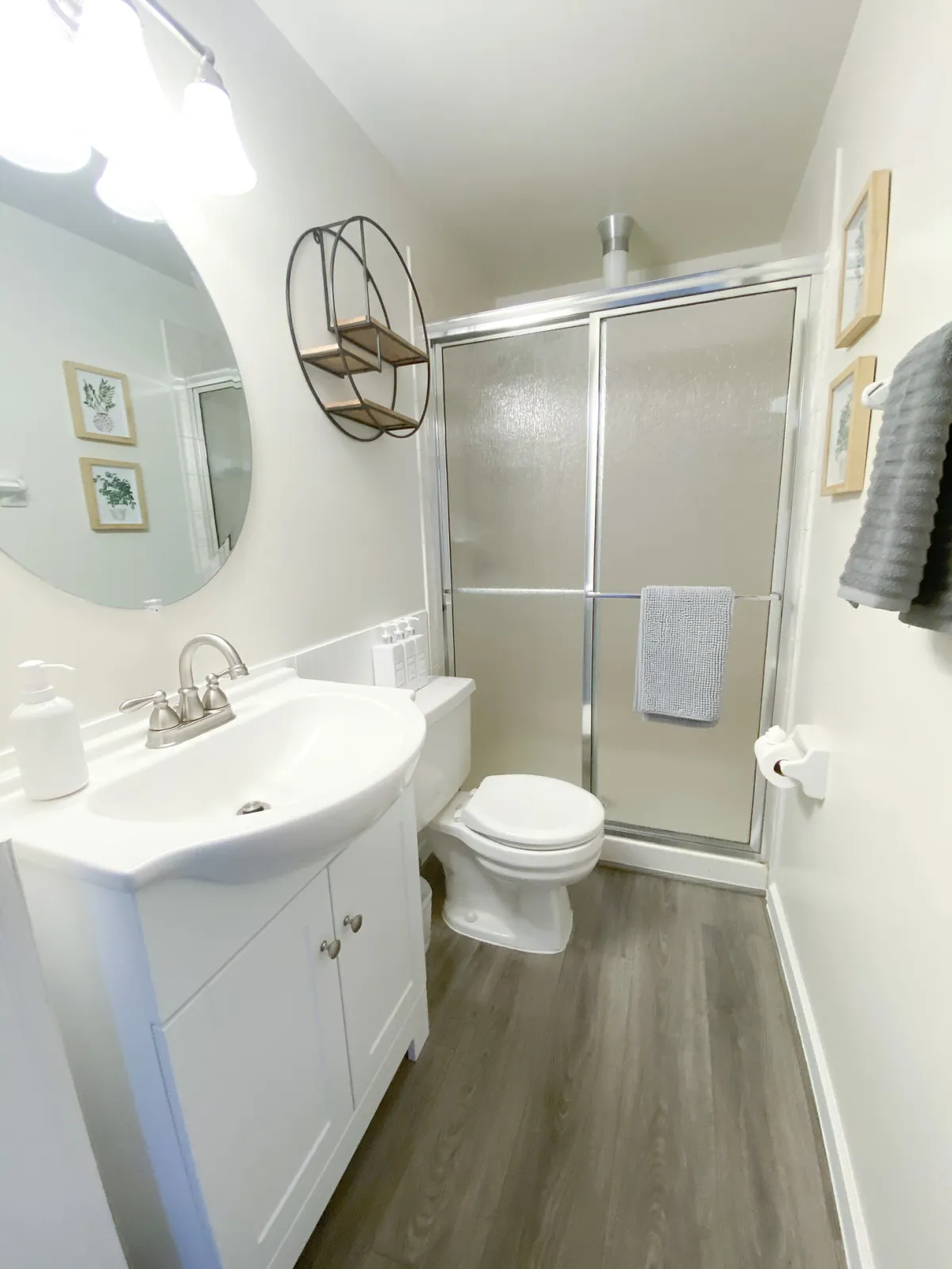 A serene bathroom with a sleek sink, glass shower, a round decorative mirror, wall shelves, and framed art. Soft morning light pours in, highlighting a space designed for rejuvenation and stress-free routines.