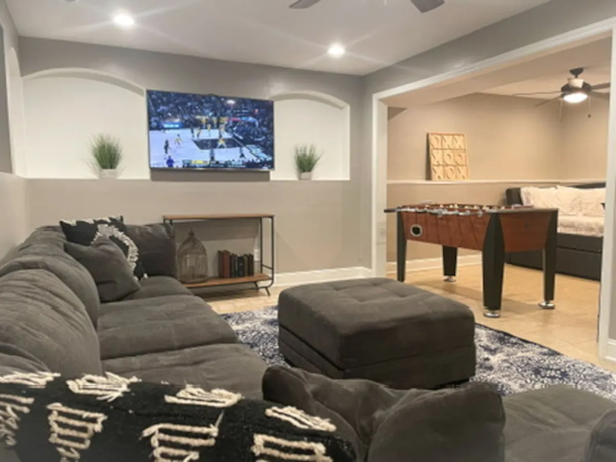 A vibrant and spacious living area with a large sectional sofa facing a television displaying a sports game, complemented by a foosball table and a decorative ottoman. A bookshelf with decorative items adds a homely touch, and the overall mood is lively and sociable.